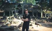 man in sunglasses with backpack stands in front of overgrown jungle temple